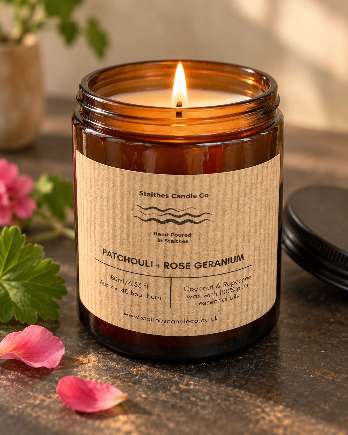 A lit amber jar with a kraft paper label reading 'Patchouli + Rose Geranium' by Stalthes Candle Co