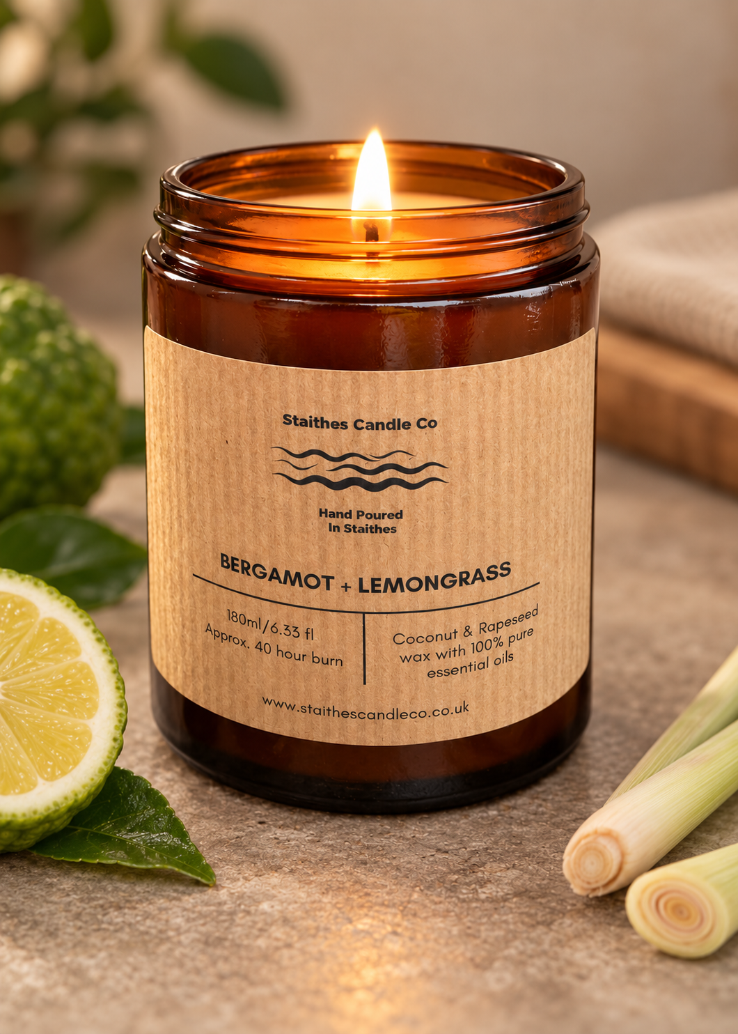 A lit amber glass candle jar with a kraft paper label reading 'Stalthes Candle Co. BERGAMOT + LEMONGRASS,' styled on a stone surface with a lime slice, green leaves, and lemongrass stems.
